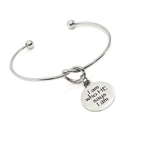Puede incluir: Un brazalete plateado con un diseño de nudo y un dije redondo. El dije está grabado con las palabras "I am who HE says I am". El brazalete tiene un diseño sencillo y elegante.