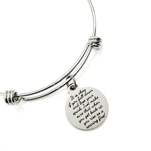 以下が含まれることがあります： 丸いチャームが付いたシルバーのバングルブレスレット。チャームには「It is okay if you fall down and lose your spark. Just make sure that when you get back up, you rise as a roaring fire!」と書かれています。