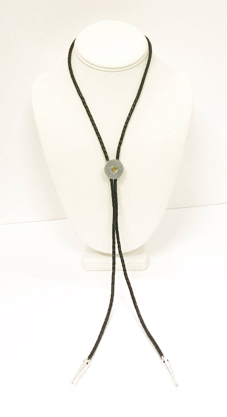 Puede incluir: Un corbat&iacute;n de cuero negro con una vaina de bala de plata como pieza central. La corbata se muestra en un maniqu&iacute; blanco.