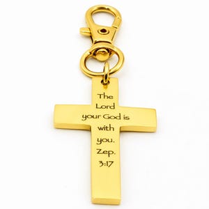 May include: A gold-coloured cross-shaped keyring with a lobster clasp. The cross features the inscription "The Lord your God is with you. Zep. 3:17". The keyring is made of a shiny, metallic material.