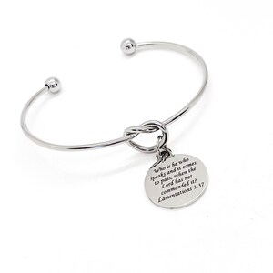 May include: Silver bangle bracelet with a knotted charm that has the quote "Who is he who speaks and it comes to pass, when the Lord has not commanded it? Lamentations 3:37" engraved on it.