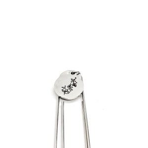 May include: Silver bookmark with a round charm featuring a floral design. The charm is attached to the bookmark with a small ring.