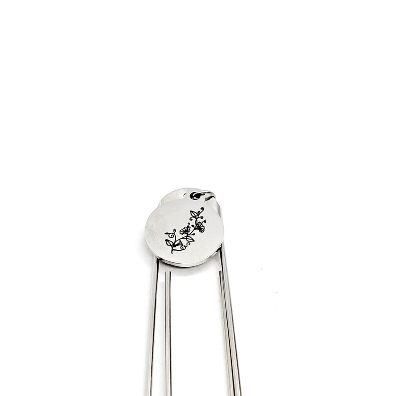 May include: Silver bookmark with a round charm featuring a floral design. The charm is attached to the bookmark with a small ring.