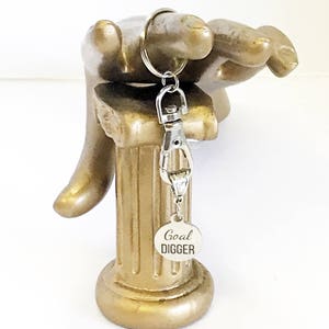 May include: Silver keychain with a round silver charm that says "Goal Digger" on a gold decorative stand.