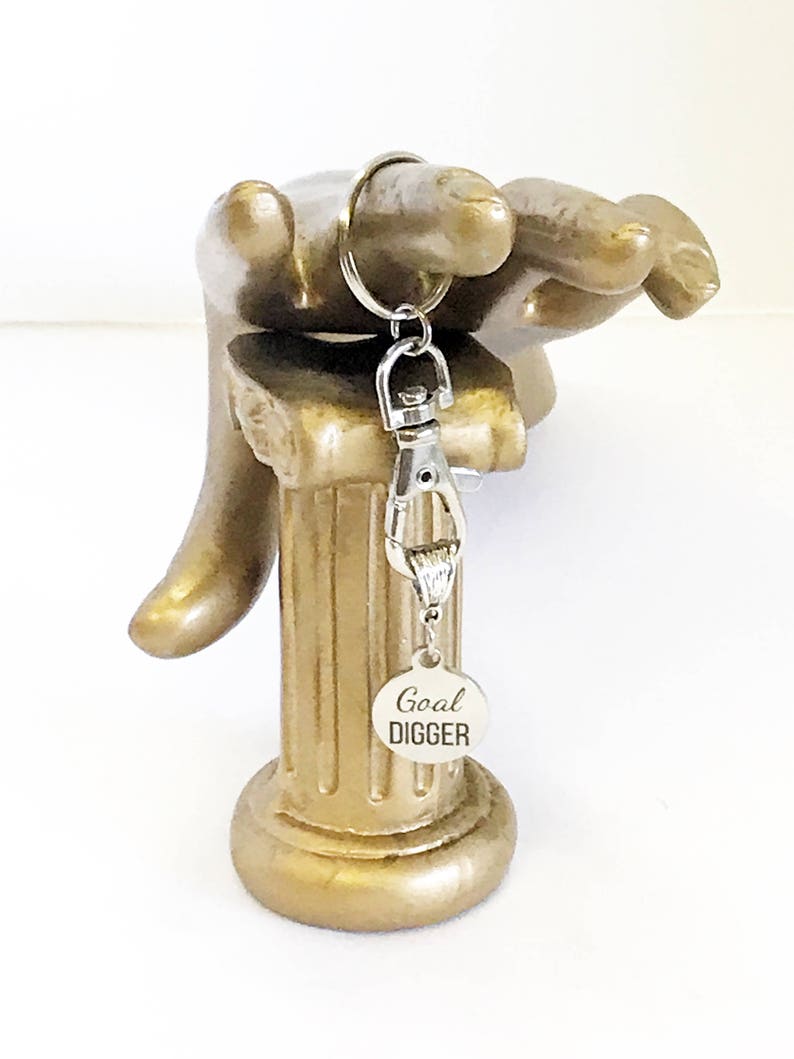 May include: Silver keychain with a round silver charm that says "Goal Digger" on a gold decorative stand.