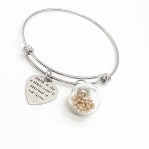 Puede incluir: Una pulsera de brazalete plateada con dos colgantes. Uno es un corazón con el texto "Faith is like a little seed if planted it will Grow". El otro es un globo de cristal lleno de pequeñas semillas beige.