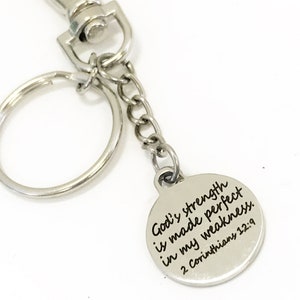 May include: Silver-toned keychain with a round charm. The charm has the inscription "God's strength is made perfect in my weakness. 2 Corinthians 12:9" in black lettering. The keychain includes a split ring, chain, and a lobster clasp.