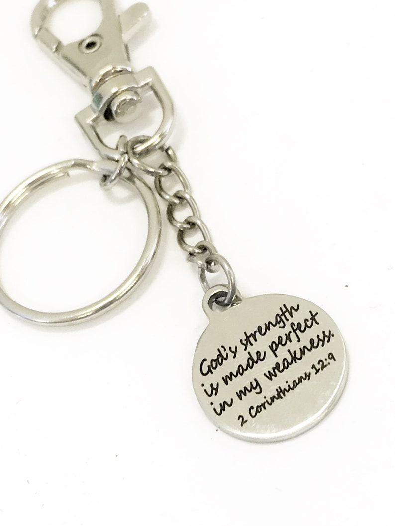 May include: Silver-toned keychain with a round charm. The charm has the inscription "God's strength is made perfect in my weakness. 2 Corinthians 12:9" in black lettering. The keychain includes a split ring, chain, and a lobster clasp.