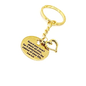 May include: Gold-tone keychain with a round charm featuring the quote "Guard your heart above all else, for it determines the course of your life. Prov 4:23" and a small heart charm.