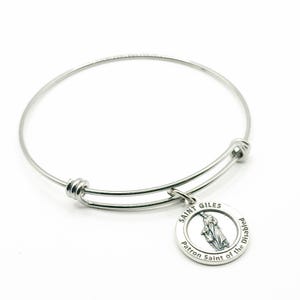 May include: A silver-colored bangle bracelet with a round charm. The charm features an image of a saint and the words "Saint Giles Patron Saint of the Disabled" around the edge. The bracelet is made of a thin, flexible metal.