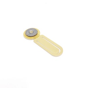 May include: A gold bookmark with a round, gray metal charm featuring a bullet casing. The charm has a gold rim and a small, gold-colored center.