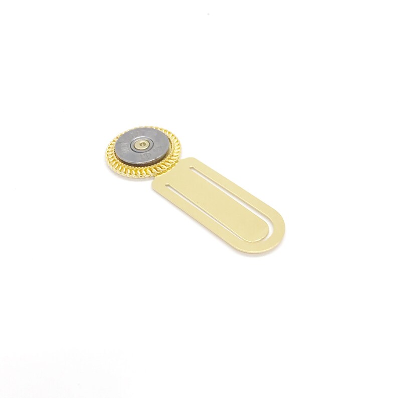 May include: A gold bookmark with a round, gray metal charm featuring a bullet casing. The charm has a gold rim and a small, gold-colored center.