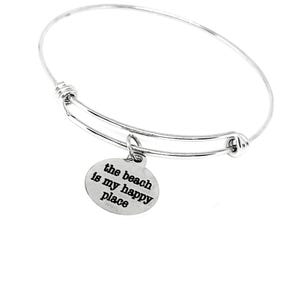 May include: A silver-colored bangle bracelet with a round charm. The charm has the phrase "the beach is my happy place" in black text. The bracelet is adjustable and has a simple, elegant design.
