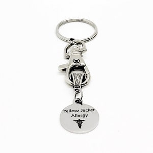 以下が含まれることがあります： 銀色のキーホルダー。銀色のクリップと、丸いタグが付いています。タグには「Yellow Jacket Allergy」と医療のシンボルが書かれています。