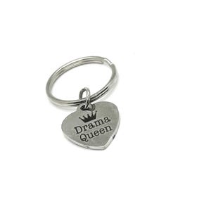 May include: A silver-colored keychain with a heart-shaped charm. The charm has the words "Drama Queen" and a crown engraved on it. The keychain is on a white background.