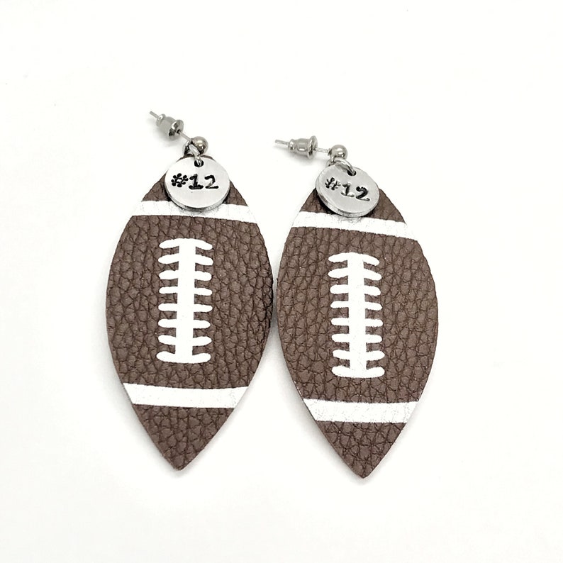 May include: A pair of brown leather football-shaped earrings with white stitching and a silver charm with the number "#12" on each earring.