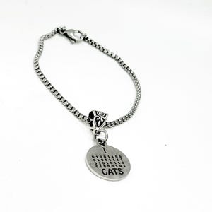 Op de afbeelding: Een zilverkleurige armband met een ronde bedel met de tekst "I CATS" en binaire code. De bedel is bevestigd aan een decoratieve kraal en een schakelarmband. De armband staat op een witte achtergrond.