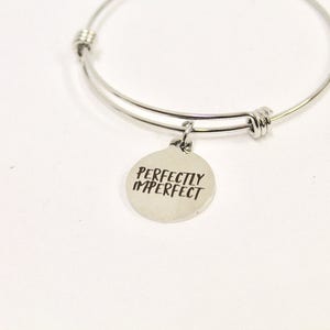 Peut inclure: Un bracelet jonc argent&eacute; avec un charme rond qui dit "Perfectly Imperfect".