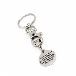 May include: A silver-colored keychain with a lobster clasp and a split ring. A small, round charm is attached, engraved with the text "Guard your heart above all else, for it determines the course of your life. Prov. 4:23."