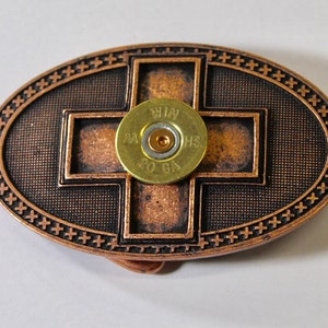 May include: A copper-toned oval belt buckle with a cross design and a gold-colored shotgun shell in the center. The buckle has a textured surface and a border of small crosses.