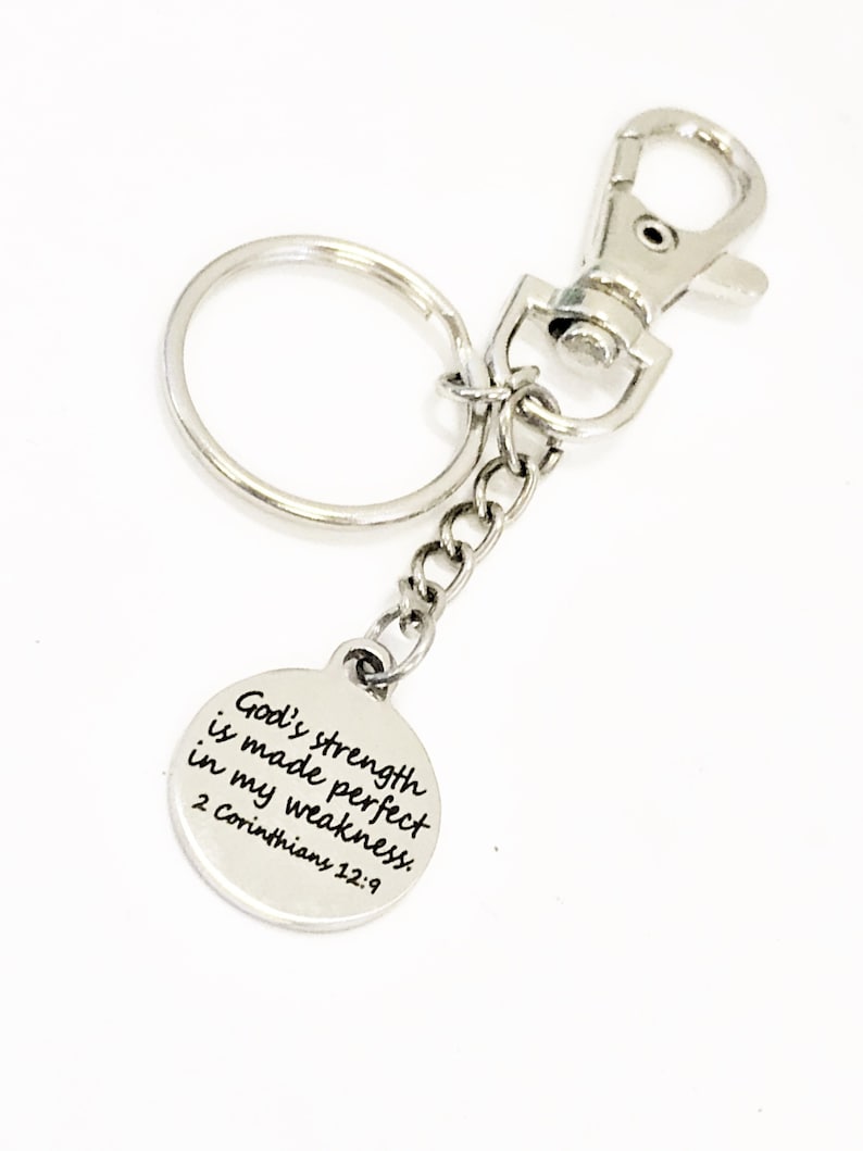 May include: A silver-colored keychain with a round charm. The charm has the text "God's strength is made perfect in my weakness. 2 Corinthians 12:9". The keychain includes a split ring, a chain, and a lobster clasp.