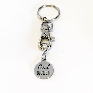 May include: Silver keychain with a silver clip and a round silver charm that says "Goal DIGGER" in black lettering.