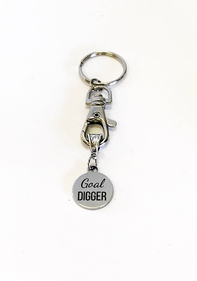 May include: Silver keychain with a silver clip and a round silver charm that says "Goal DIGGER" in black lettering.