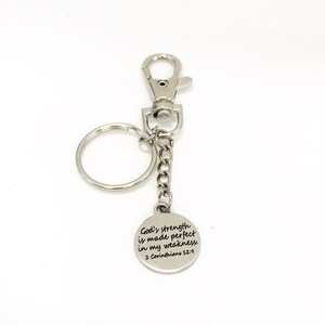 May include: A silver-colored keychain with a round charm. The charm has the text "God's strength is made perfect in my weakness 2 Corinthians 12:9". The keychain includes a clasp and a key ring.
