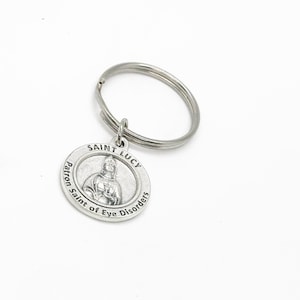 May include: Silver keychain with a round charm featuring a depiction of Saint Lucy, the patron saint of eye disorders. The charm has the words "Saint Lucy" and "Patron Saint of Eye Disorders" inscribed around the image.