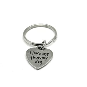 May include: A silver-colored keychain with a heart-shaped charm. The charm has the phrase "I love my therapy dog" in a handwritten style font. The keychain is on a white background.