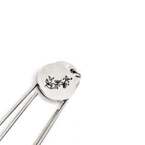 May include: Silver bookmark with a floral design engraved on a round charm. The bookmark is made of metal and has a simple, elegant design.