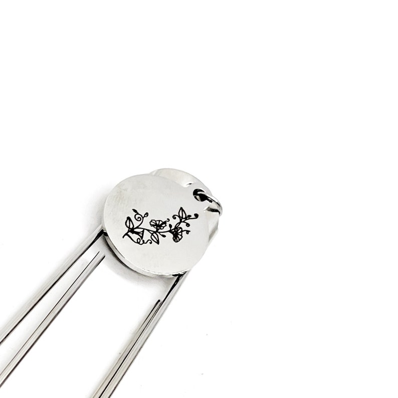 May include: Silver bookmark with a floral design engraved on a round charm. The bookmark is made of metal and has a simple, elegant design.