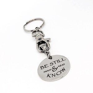 May include: A silver-colored keychain with a round pendant. The pendant has the words "BE STILL & KNOW" and "PSALM 46:10" engraved on it. The keychain includes a clasp and a key ring.