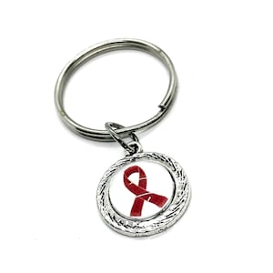 May include: Silver-toned keychain with a round charm featuring a red ribbon design on a white background. The charm has a textured, braided border. The keychain is on a white background.