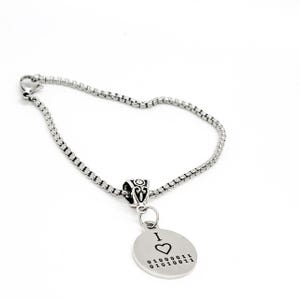 May include: A silver chain bracelet with a silver charm pendant. The charm is round and has the text "I love 01000011 01010011" engraved on it.