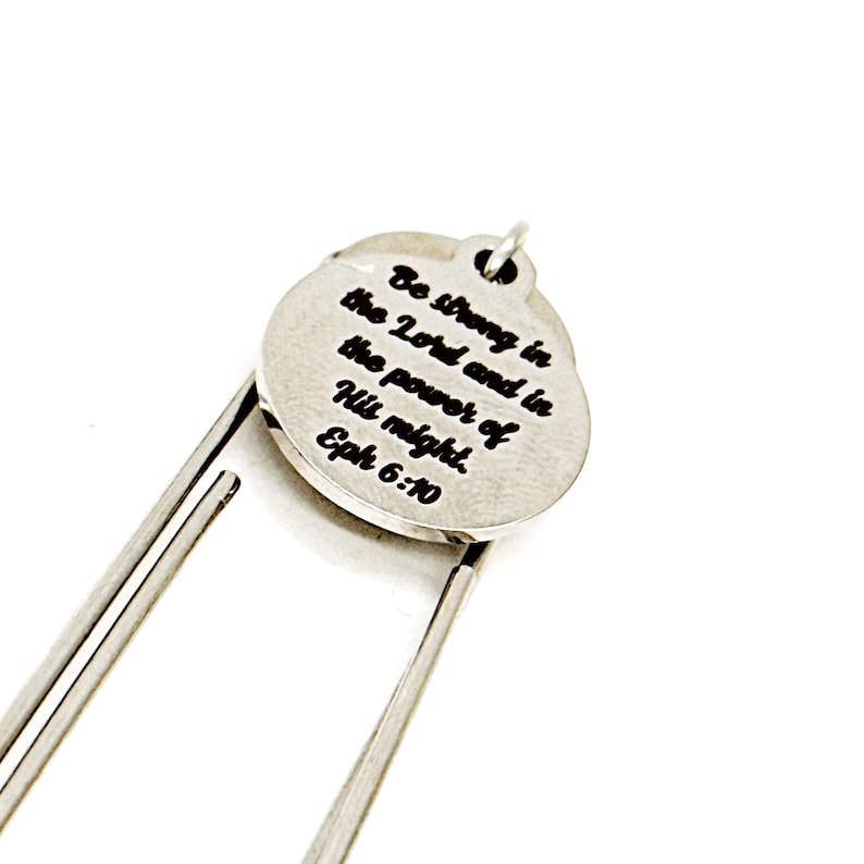 Puede incluir: Marcap&aacute;ginas de plata con un encanto redondo que dice "Sed fuertes en el Se&ntilde;or y en el poder de su fuerza. Efesios 6:10".