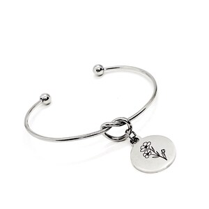 May include: Silver knot bangle bracelet with a round charm featuring a delicate floral design.