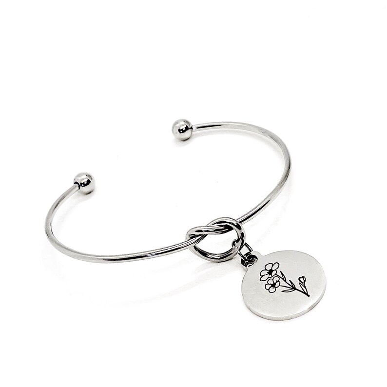 May include: Silver knot bangle bracelet with a round charm featuring a delicate floral design.