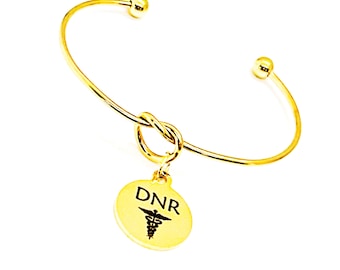 May include: Gold bangle bracelet with a knot detail and a round charm with the text "DNR" and a medical symbol.