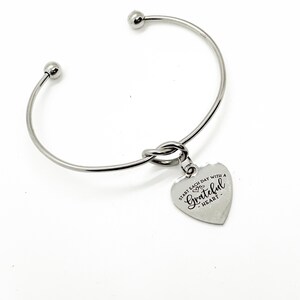 May include: Silver bangle bracelet with a knotted design and a heart-shaped charm that reads "Start each day with a grateful heart".