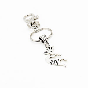 May include: A silver key chain with a Kokopelli charm. The charm is a stylized figure of a flute-playing man with a headdress.