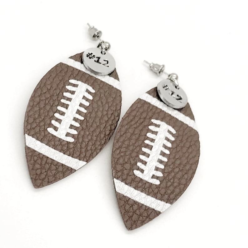 May include: A pair of brown leather football-shaped earrings with white stitching and a silver charm with the number "#12" on each earring.