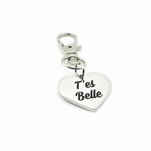 以下が含まれることがあります： ハート型のチャームが付いたシルバーカラーのキーホルダー。「T'es Belle」というフレーズが黒い筆記体で書かれています。キーホルダーには、鍵やバッグに簡単に取り付けられる留め金が付いています。