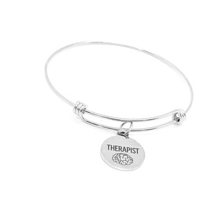 May include: A silver-colored bangle bracelet with a round charm. The charm is engraved with the word "THERAPIST" and a brain graphic. The bracelet is made of a thin, flexible metal and has an adjustable design.