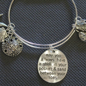 Może przedstawiać: Srebrna bransoletka z zawieszką z cytatem "may you always have a shell in your pocket & sand between your toes". Bransoletka ozdobiona jest zawieszkami w kształcie gwiazd morskich, muszli, piaskowego dolara i muszli nautilusa.