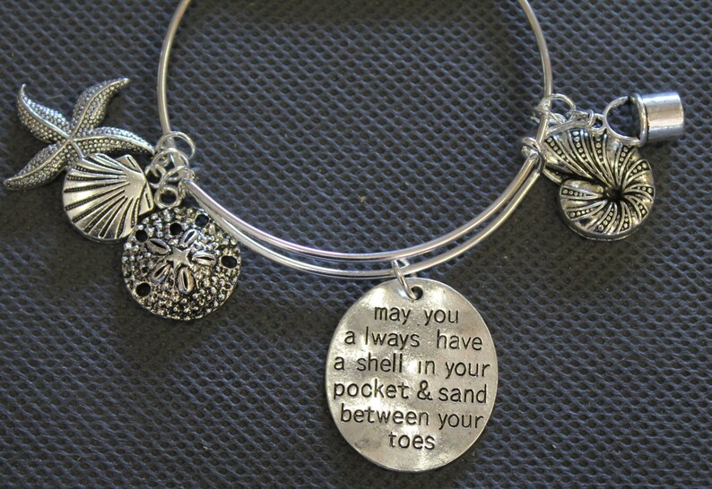 Może przedstawiać: Srebrna bransoletka z zawieszką z cytatem "may you always have a shell in your pocket & sand between your toes". Bransoletka ozdobiona jest zawieszkami w kształcie gwiazd morskich, muszli, piaskowego dolara i muszli nautilusa.