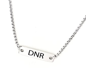 May include: A silver necklace featuring a rectangular pendant with the letters "DNR" in black. The chain has a fine, linked pattern. The necklace is shown against a white backdrop.