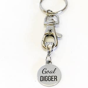 May include: Silver keychain with a silver clip and a round silver charm that says "Goal DIGGER" in black lettering.