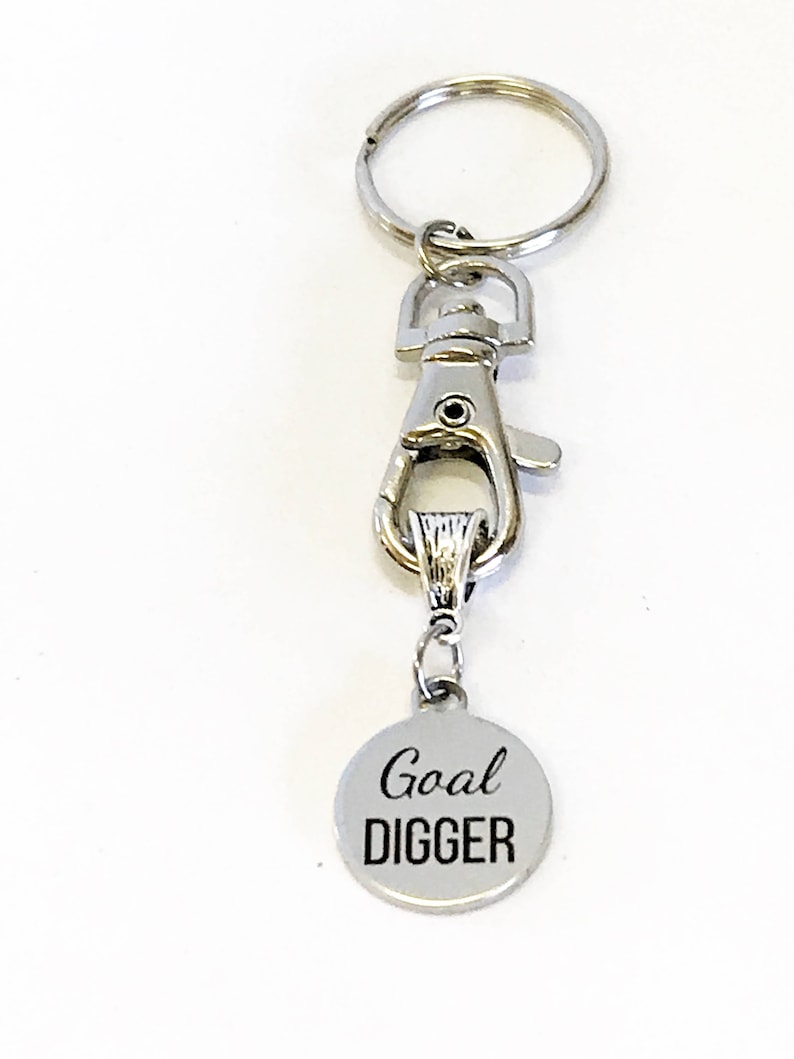 May include: Silver keychain with a silver clip and a round silver charm that says "Goal DIGGER" in black lettering.