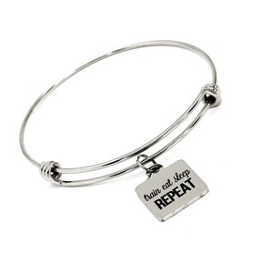 May include: Silver expandable bangle bracelet with a square charm that reads "train eat sleep repeat" in black lettering.